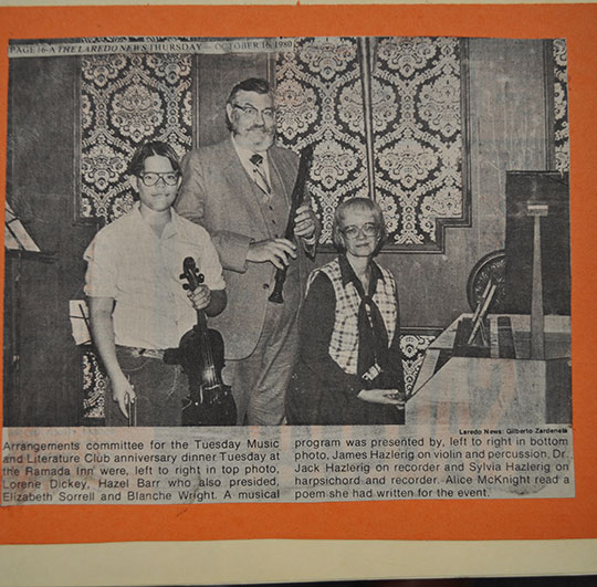 A young boy with oversized glasses and medium width hair that is parted down the middle is holding a violin. Next to him are his parents. His father, who sports a grand beard, holds a recorder while his mother, a woman with short hair fashioned into a bob cut, is seated at a piano.