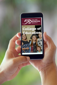Stock photo of a person's hand holding a smartphone. The phone is displaying the TAMIU College of Education homepage.
