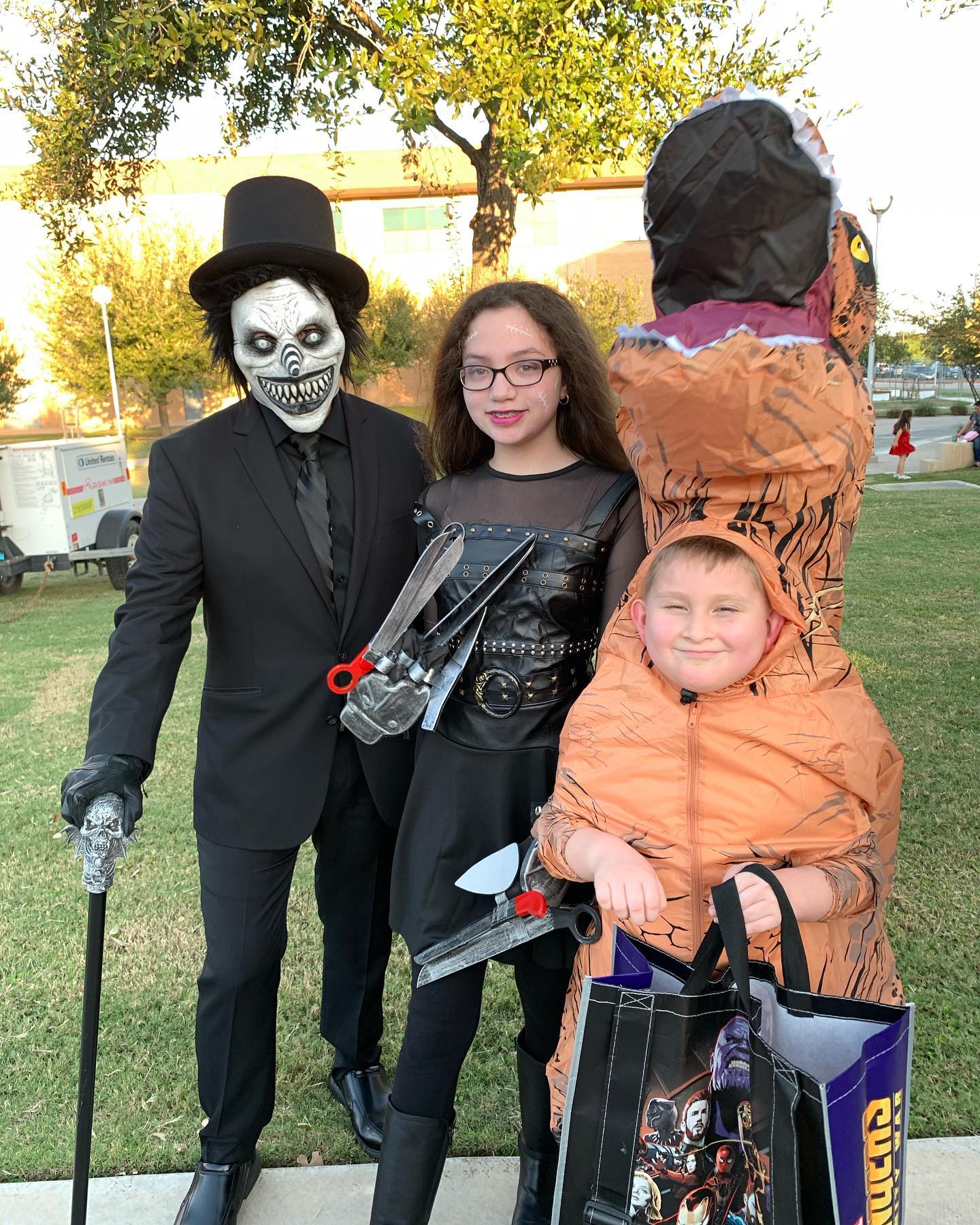 Three individuals in halloween costumes: a masked person with a suit, a young female with scissor hands, a young boy with an inflatable tyrannosaurus rex costume